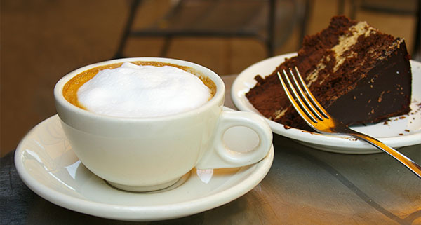 a cup of frothy coffee next to a plate of a chocolate cake with a fork